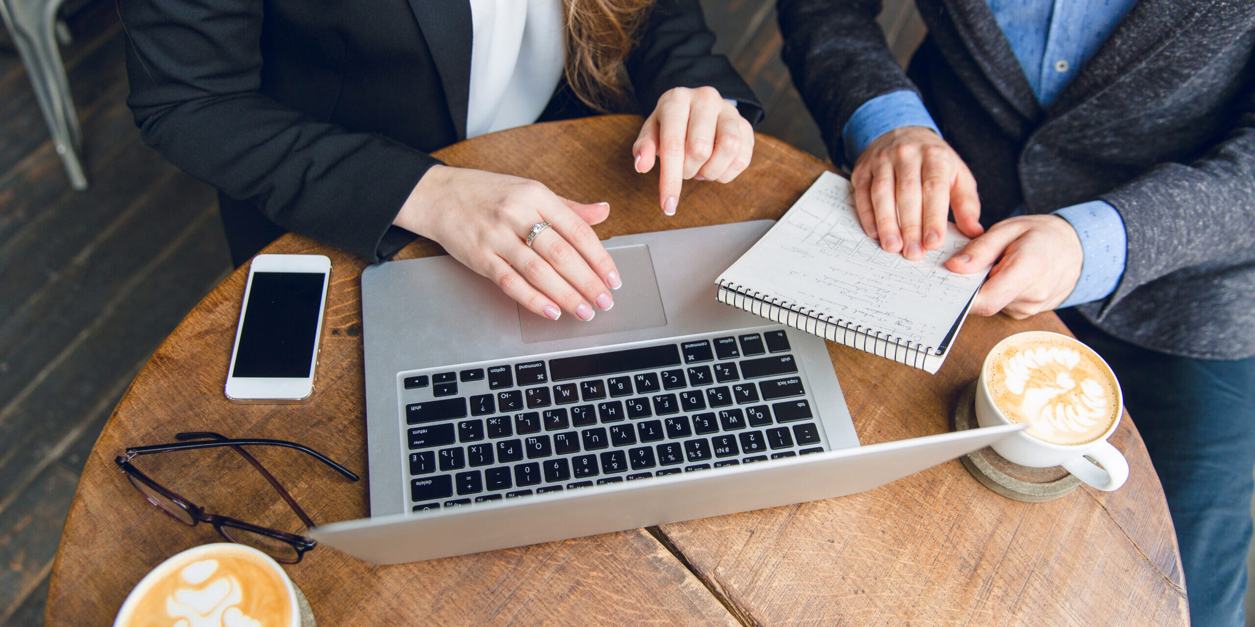 close-up-coffee-table-with-two-colleagues-sitting-holding-notebook-typing-laptop