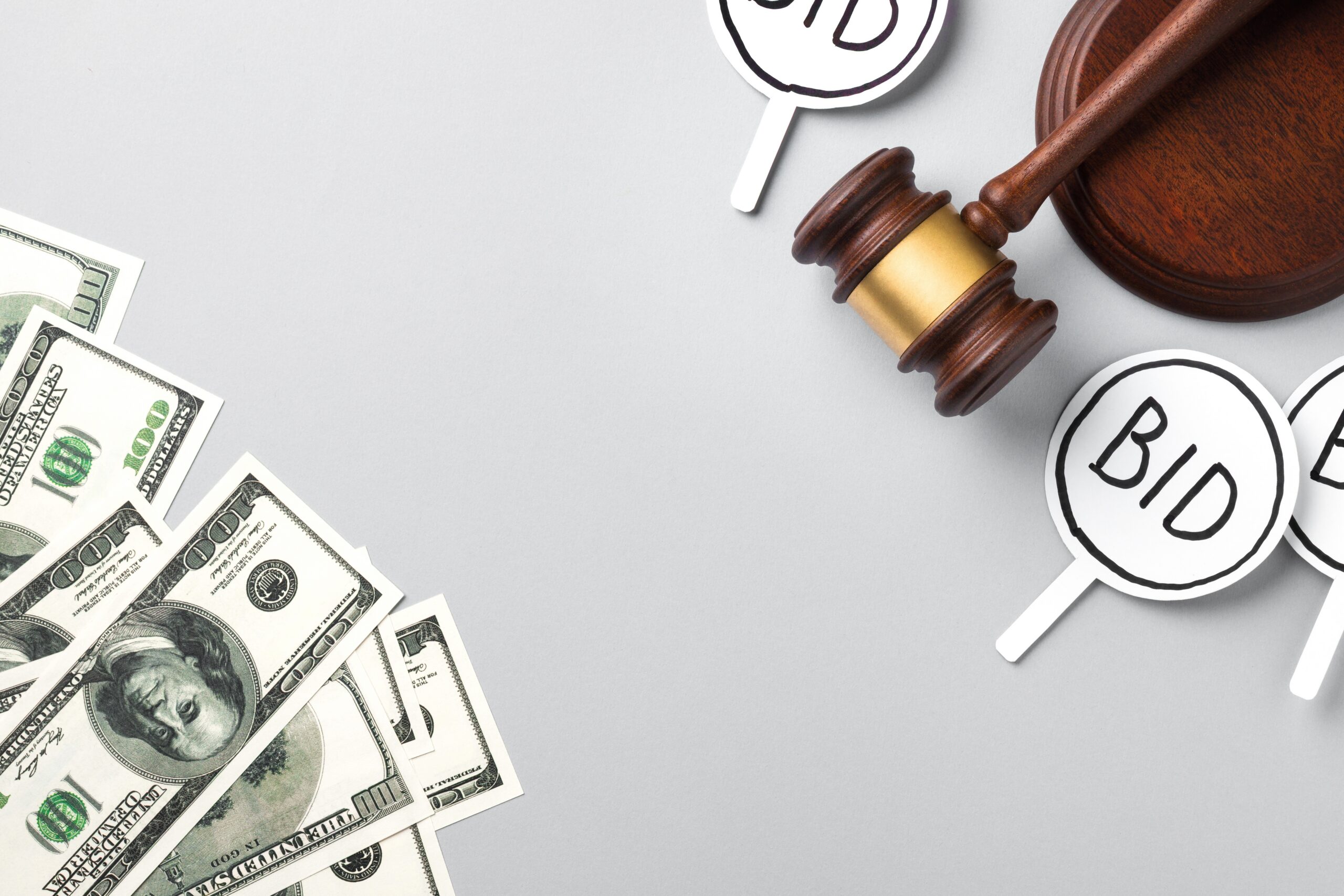 A wooden auction gavel, white bidding paddles with "BID" signs and US dollar bills on a light gray surface. Concept of auction, bidding, money, finance and sales. Top view with copy space.